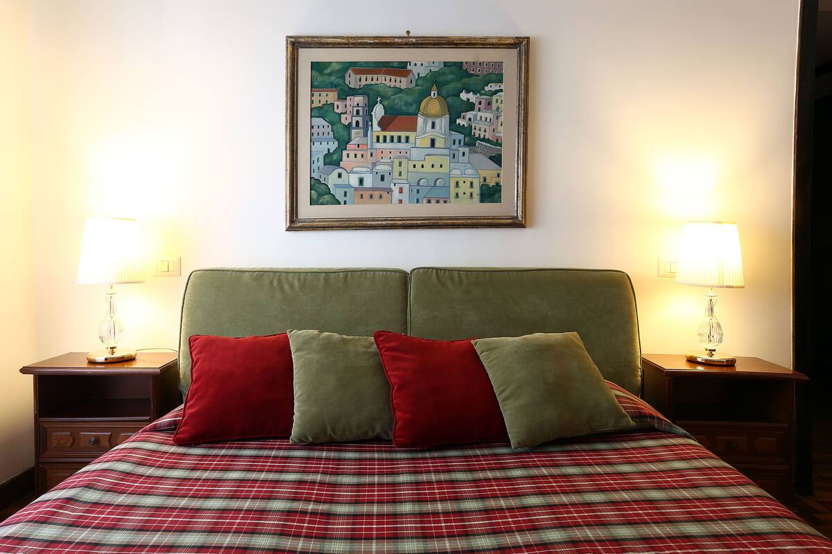 A neatly made bed with plaid bedding, two bedside lamps, and a framed cityscape painting above it.