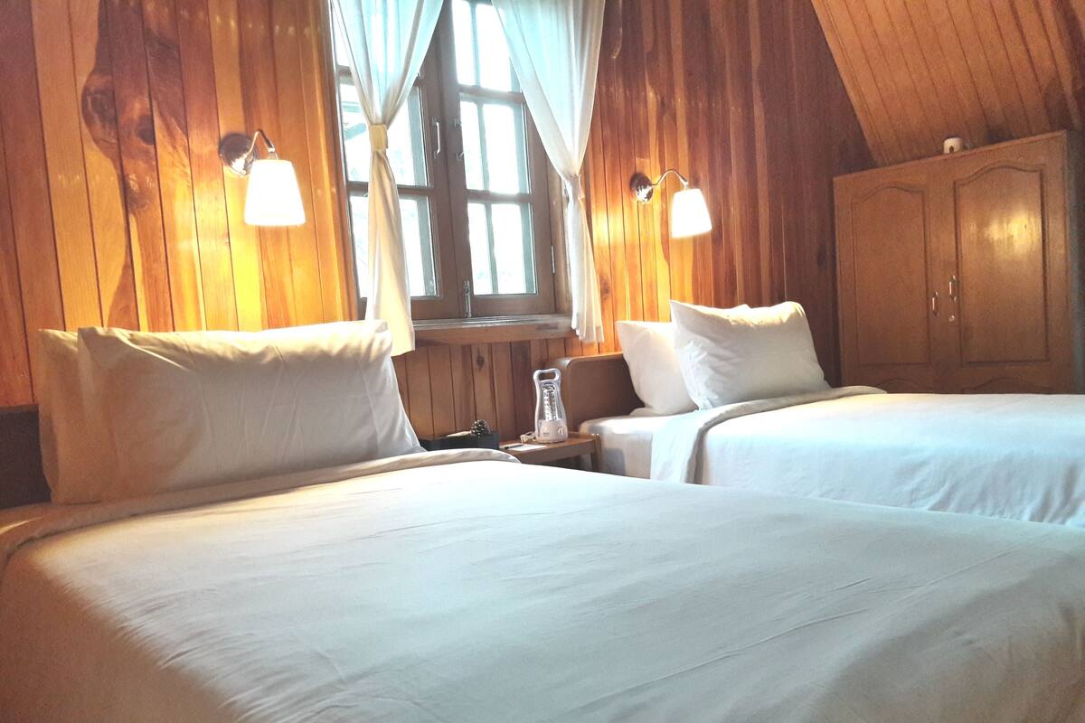 A room with two beds, wooden walls, a window with curtains, and a wooden wardrobe.