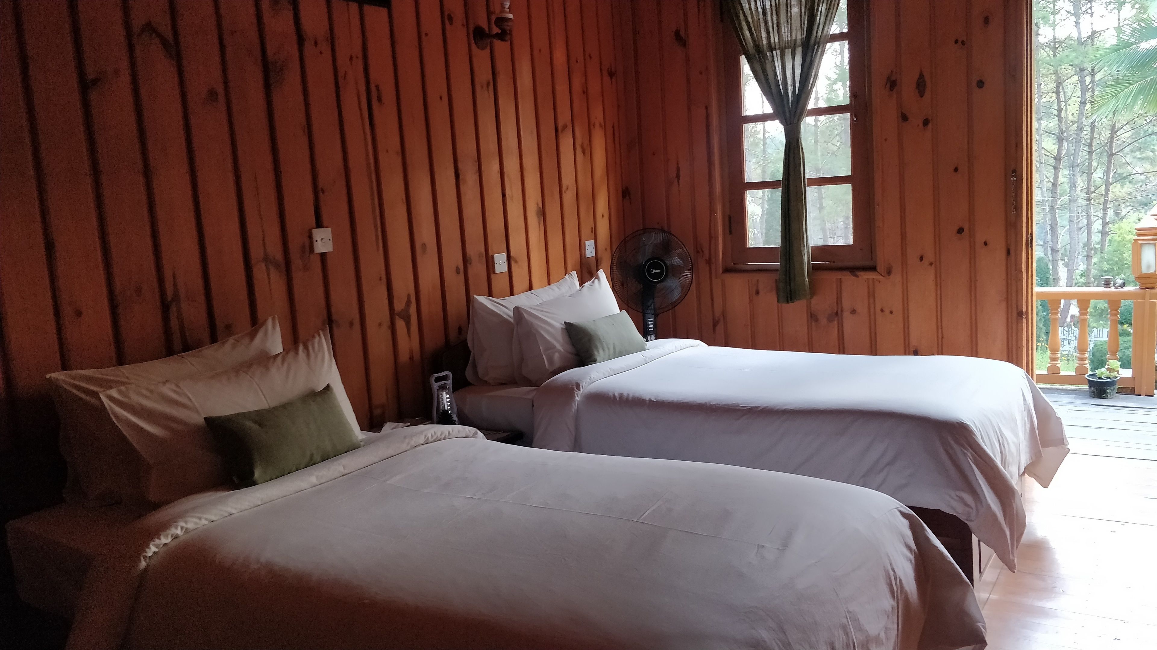 Una habitación tipo cabaña con dos camas, paredes de madera, una ventana con vistas a árboles y un ventilador en la pared.