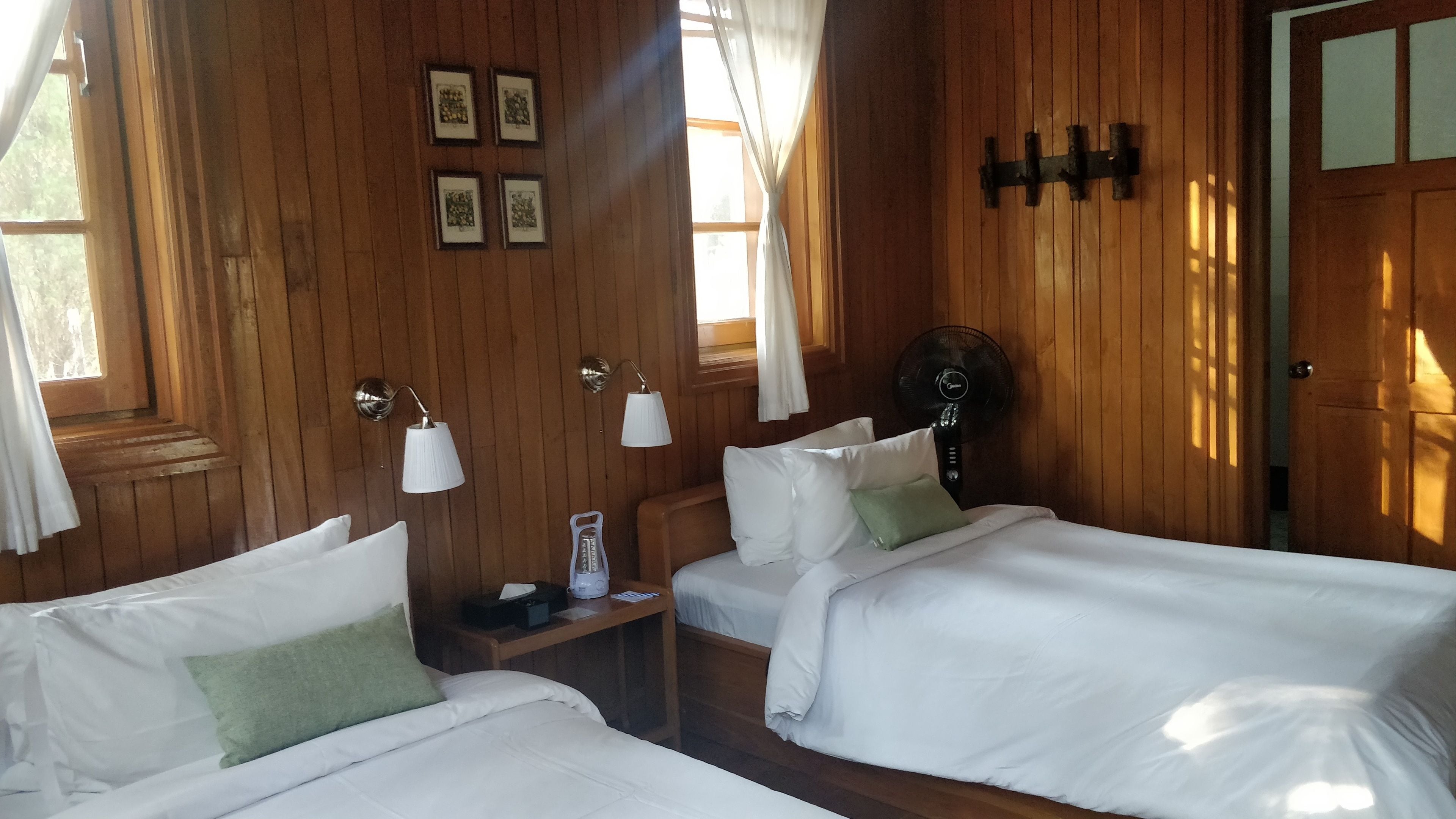 A wooden room with two beds, a nightstand, a lamp, and a fan.