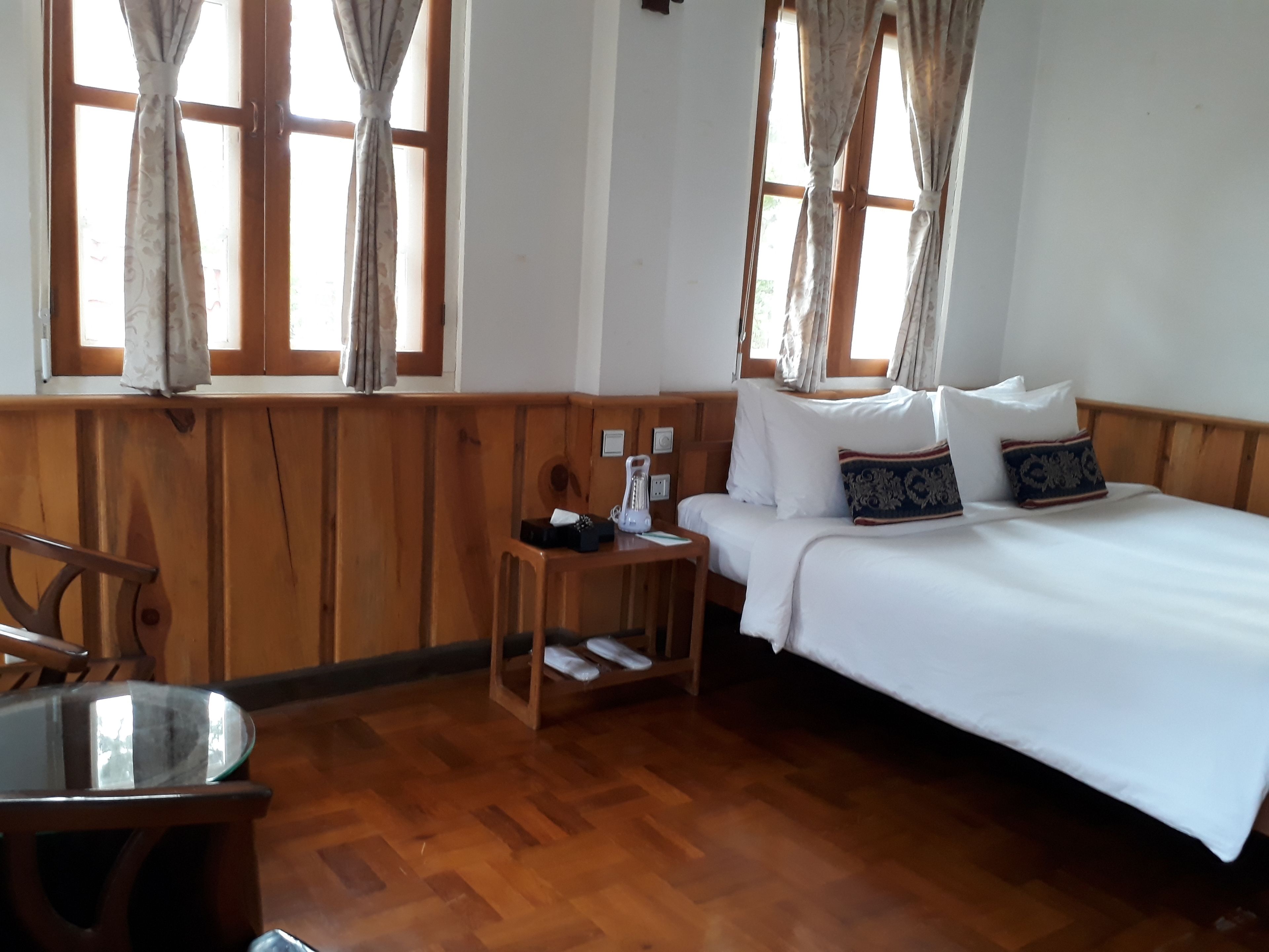 A room with a wooden floor, a bed with white bedding and decorative pillows, a wooden nightstand with a lamp, and a window with curtains.