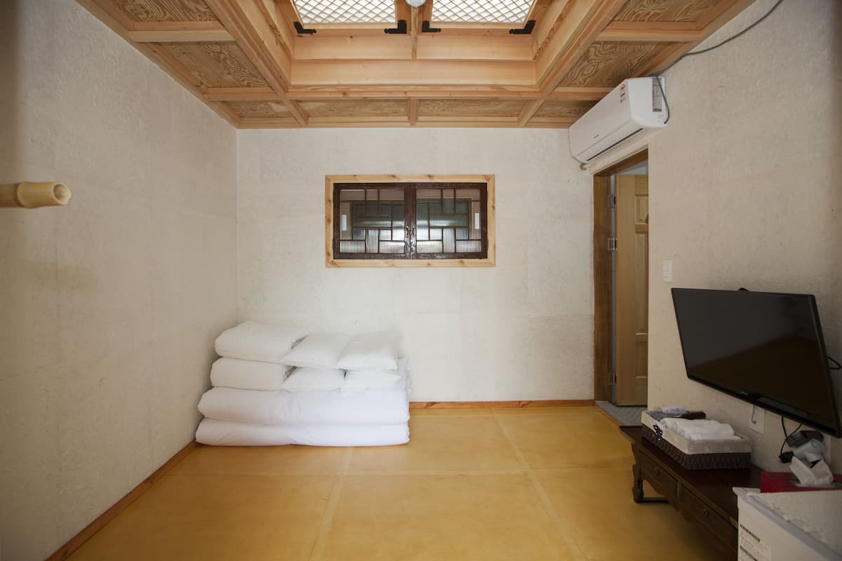 A room with a wooden ceiling, a window with a traditional design, a stack of white towels, and a flat-screen TV on a wooden stand.