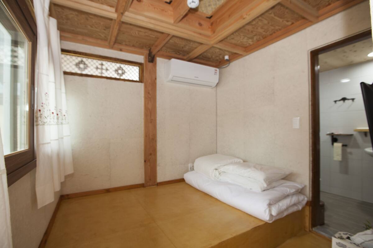 A room with a wooden ceiling, a bed with white bedding, an air conditioning unit, and a window with a decorative screen.
