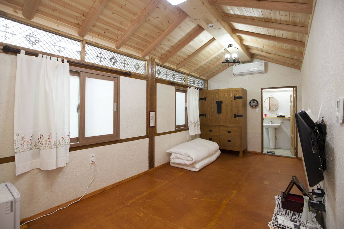 A room with wooden beams, a large window with curtains, a wooden cabinet, a sink, and a computer monitor.