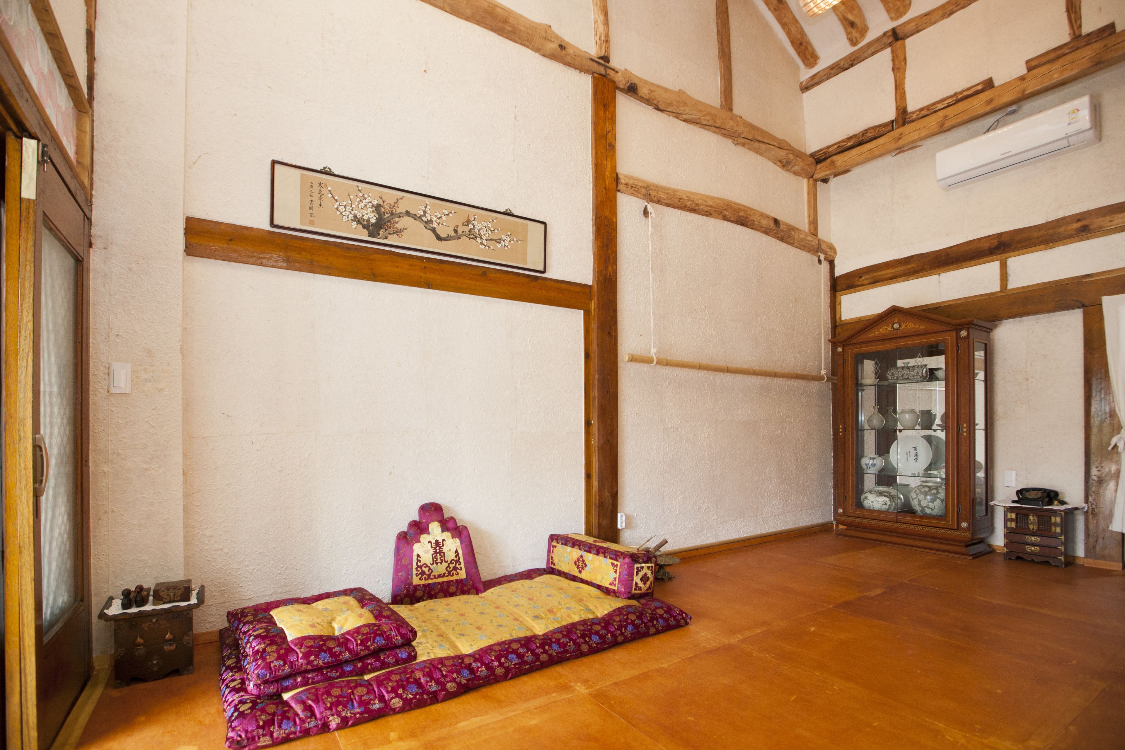 A traditional Japanese-style room with tatami flooring, a low wooden bed with a purple and yellow bedspread, a wooden cabinet with glass doors, and a framed artwork on the wall.