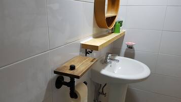 Un baño con un lavamanos blanco, un estante de madera, un espejo y un dispensador de papel higiénico.