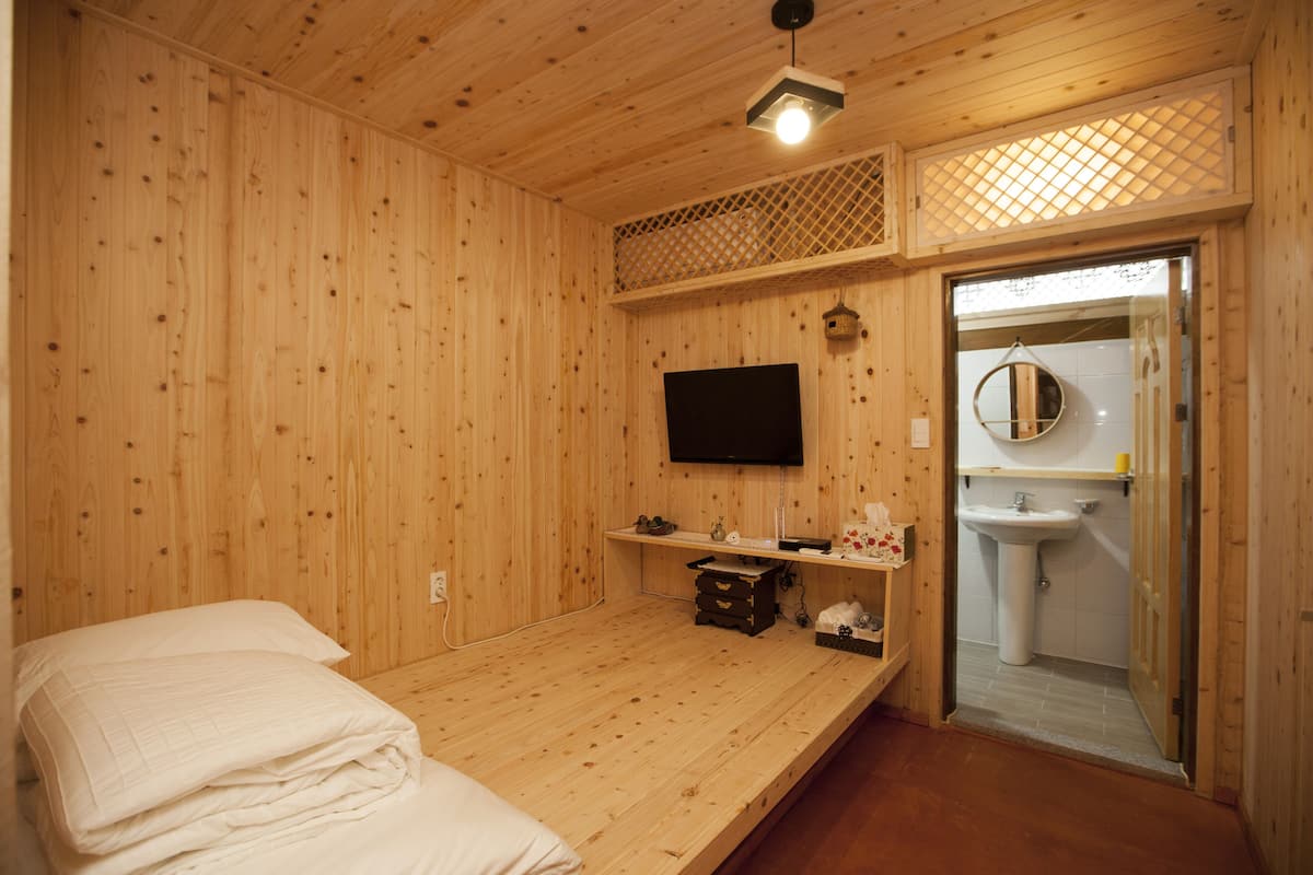 A wooden room with a bed, a television, a shelf with toiletries, and a bathroom visible through an open door.