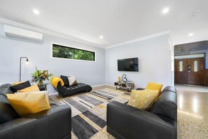 Een moderne woonkamer met twee leren zwartsofa’s, een kamerbreed tapijt met dessin, een flatscreen televisie en een raam met uitzicht op groen.