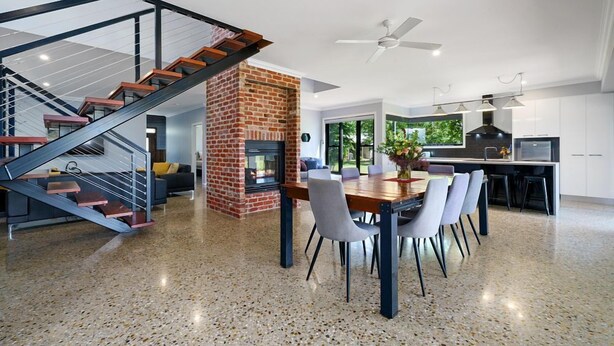 A modern kitchen with a central island, a dining area with chairs, a brick fireplace, and a staircase with glass railings.