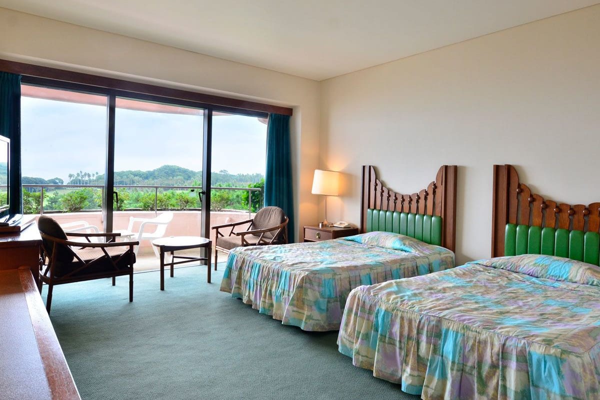 Une chambre d’hôtel avec deux lits, un bureau et une chaise. Elle dispose d’un balcon avec vue sur les collines.