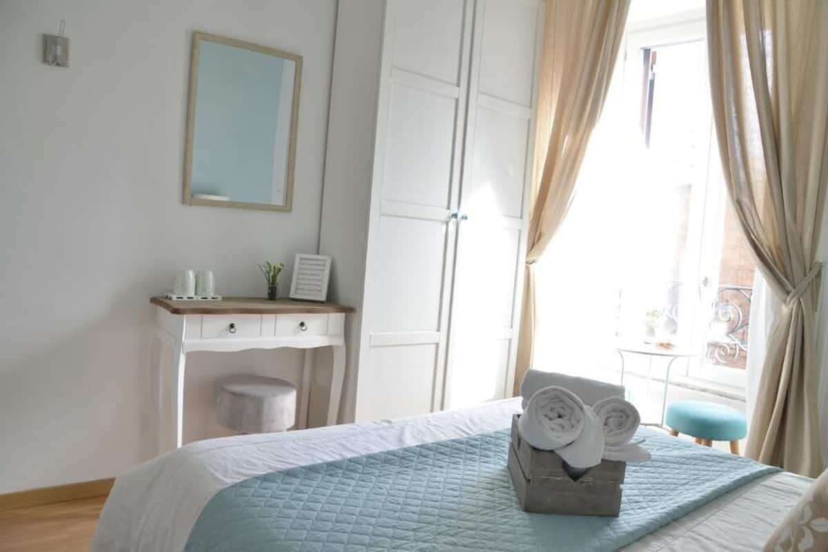 Une chambre d’hôtel comprenant un lit, une table de chevet avec une lampe, un miroir, une armoire et une fenêtre avec des rideaux.