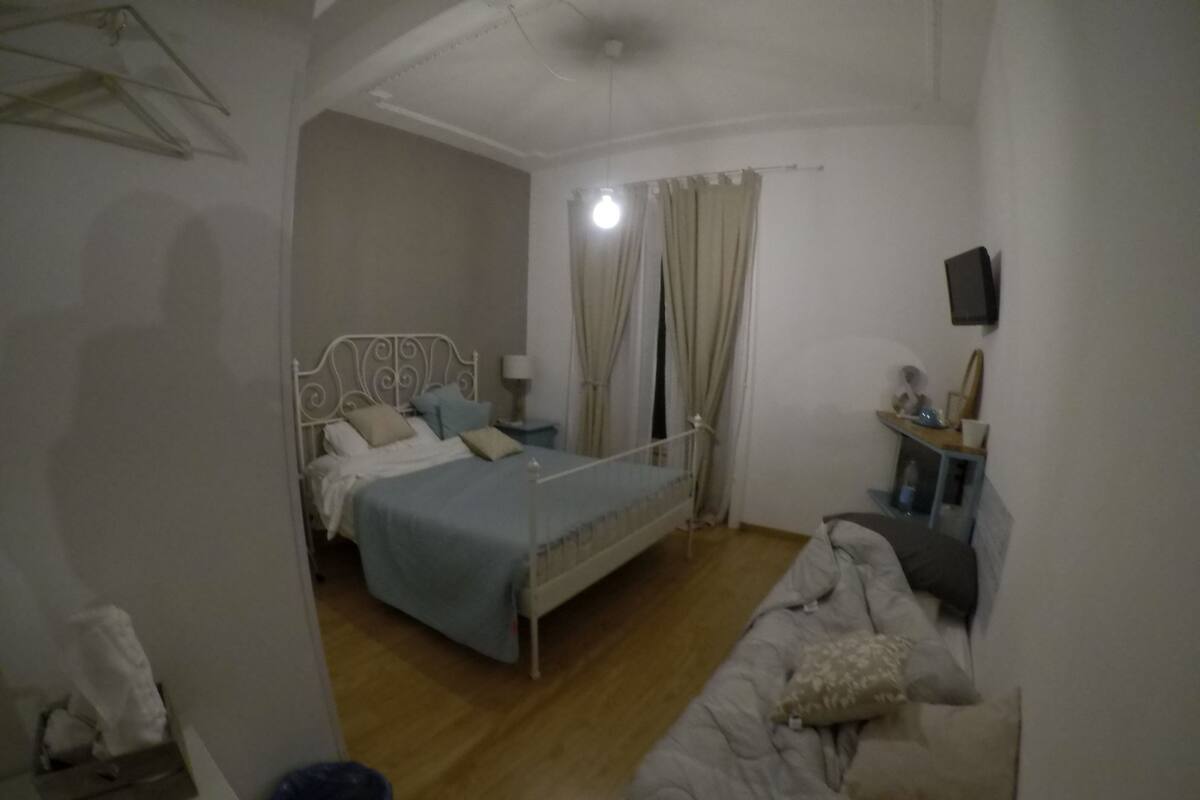 Une chambre avec un lit simple, une table de chevet, une chaise, un téléviseur fixé au mur et une fenêtre avec des rideaux.