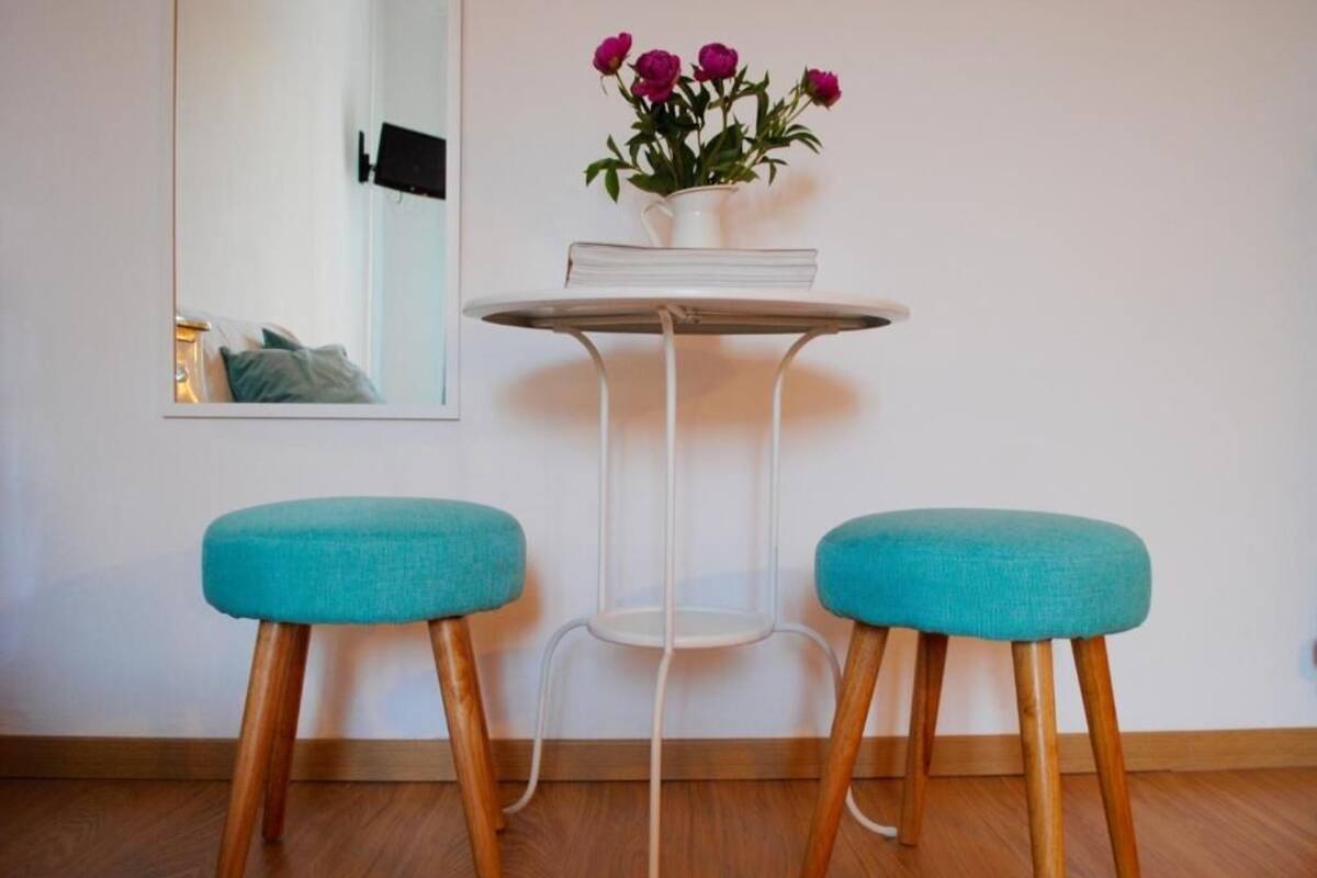 Une pièce avec un plancher en bois, un mur blanc, une table ronde avec une chaise blanche, deux tabourets rembourrés de couleur turquoise et un vase avec des fleurs sur la table.
