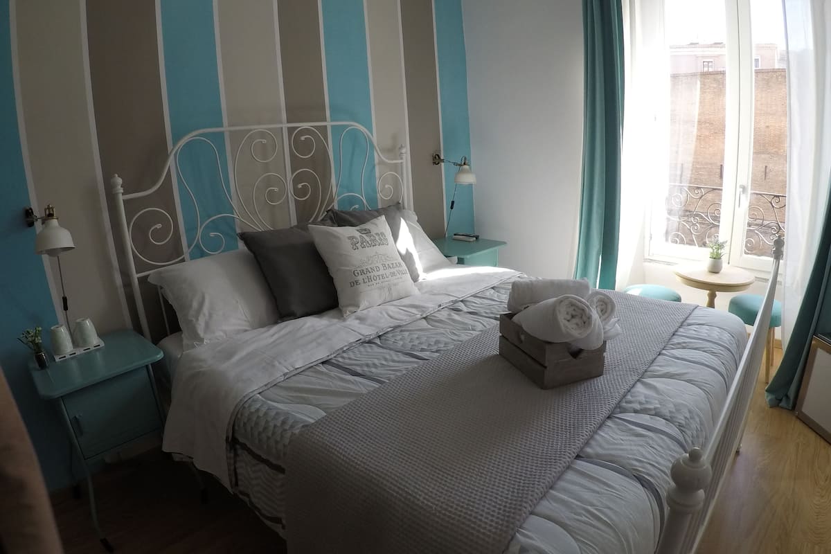 Un lit bien fait, avec une couette blanche et des oreillers, une table de chevet avec une lampe, et une vue sur l’extérieur par une fenêtre.