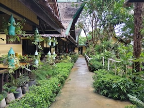 A narrow pathway flanked by lush greenery and potted plants, leading to a building with a thatched roof and wooden beams. - Cliffside Cottages (El Nido)