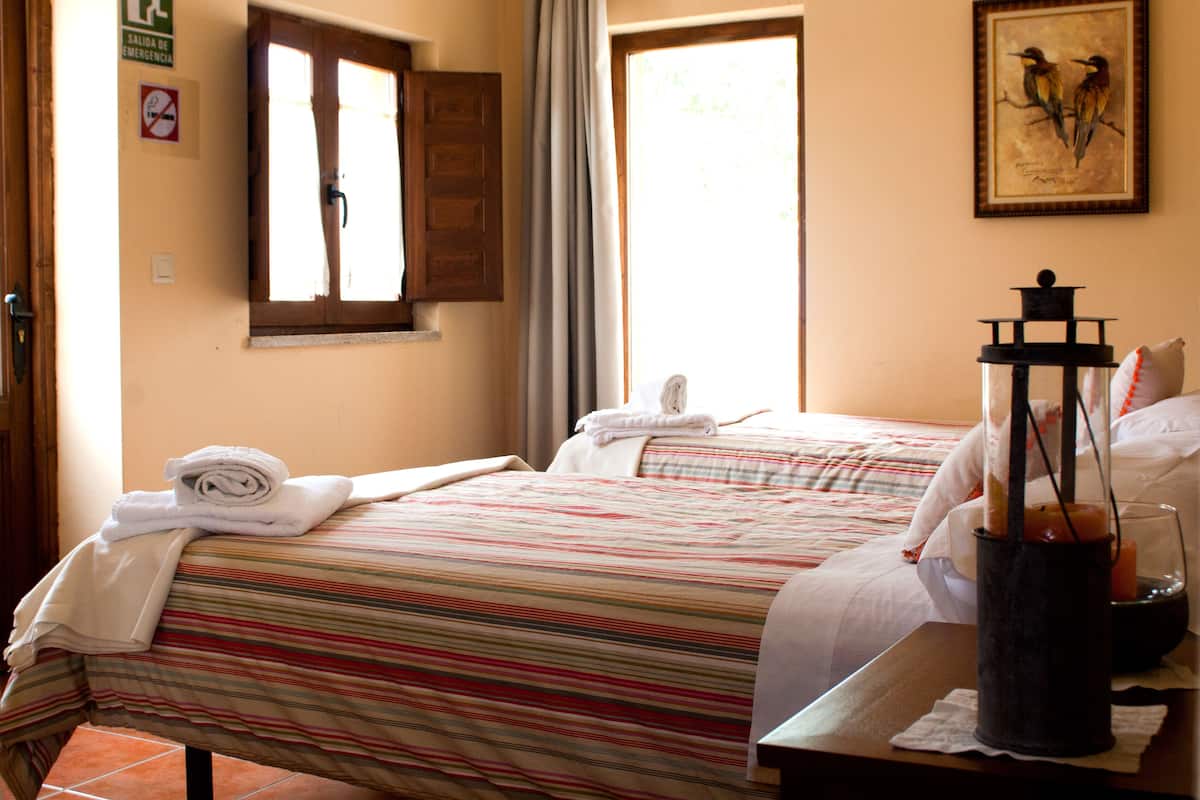 A hotel room with two beds, a window with curtains, a framed bird painting, and a sign indicating an emergency exit.