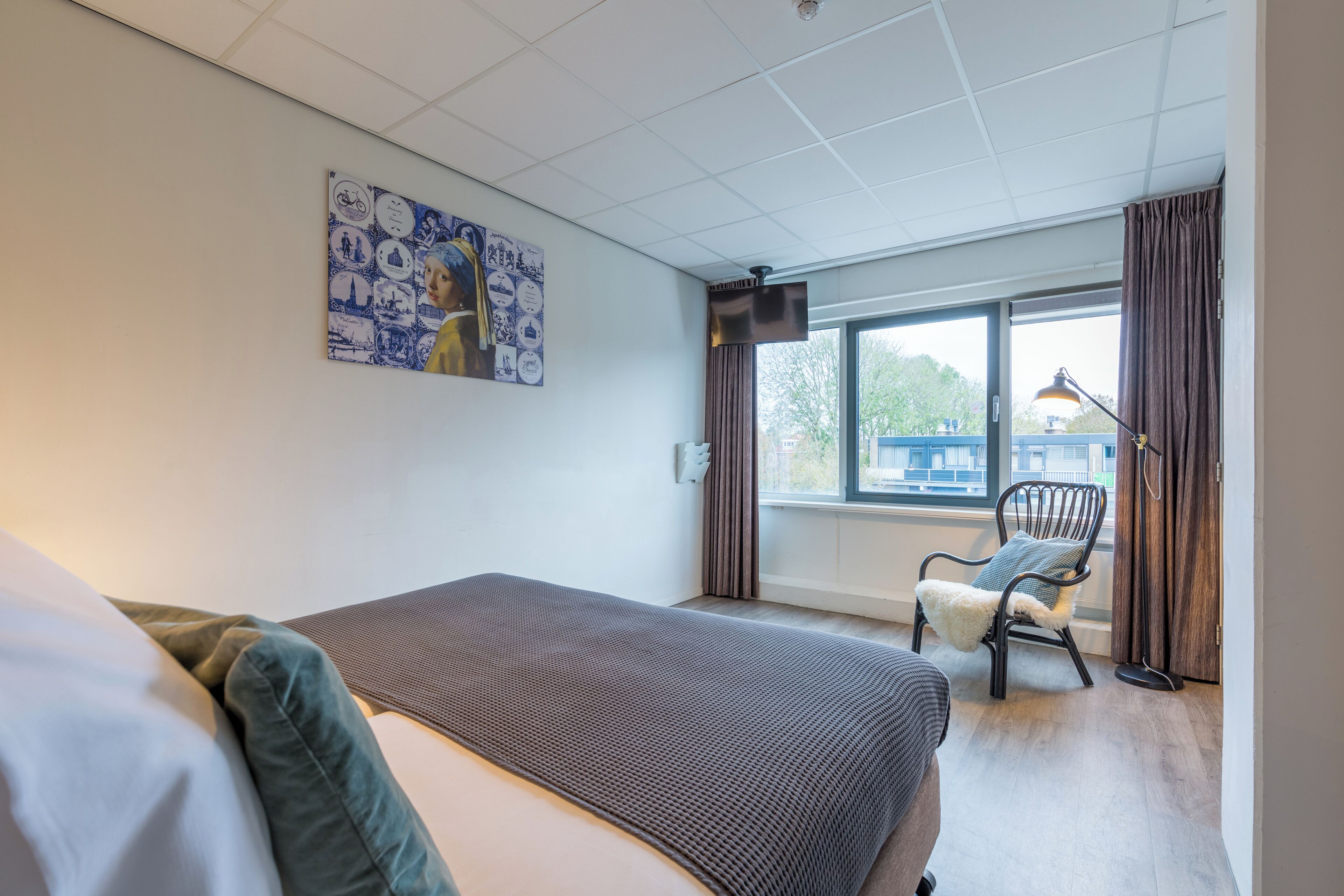 Une chambre d’hôtel avec un lit, une chaise, une fenêtre avec des rideaux et un mur orné d’une affiche.