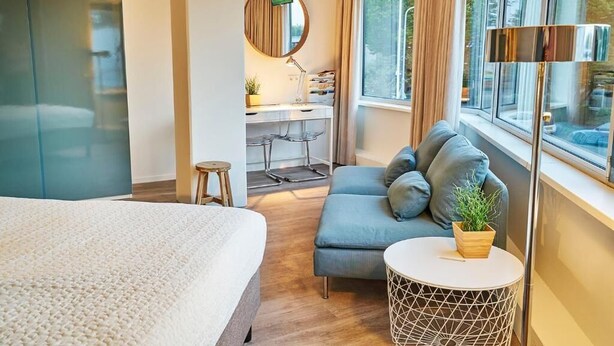Ein modernes Hotelzimmer mit einem großen Bett, einer Sitzecke, einem kleinen runden Tisch, einem Schreibtisch mit Stuhl, einem Spiegel und einem Fenster mit Vorhängen.