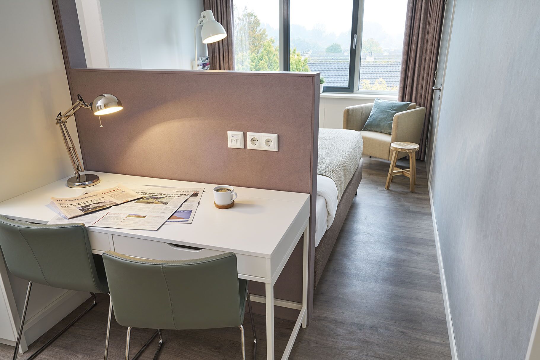 A hotel room with a desk, two chairs, a bed, a window with curtains, and a bedside table with a lamp and a cup.