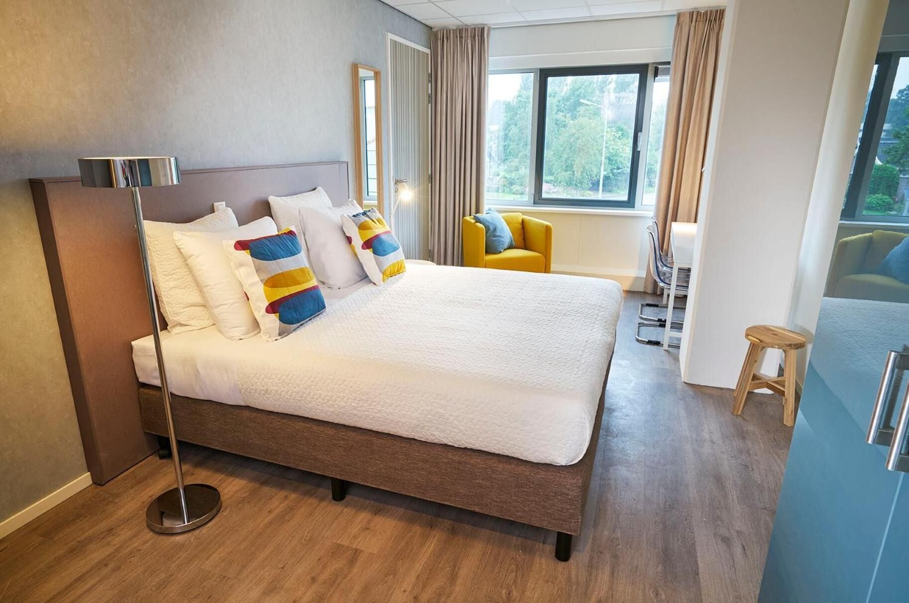 A modern hotel room with a large bed, a bedside lamp, a chair, and a view of greenery outside.