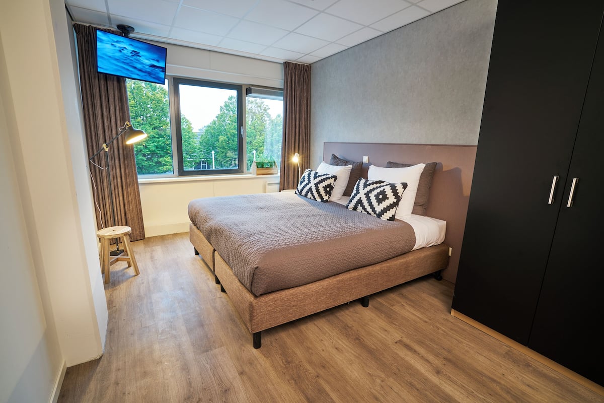 Un dormitorio moderno con una cama grande, un televisor en la pared, una ventana con cortinas y un armario.