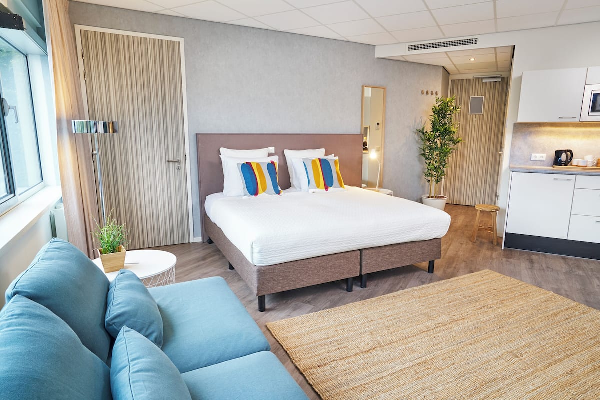 Una habitación de hotel moderna con una cama grande, un sofá, una pequeña mesa redonda, una planta en maceta y una kitchenette.