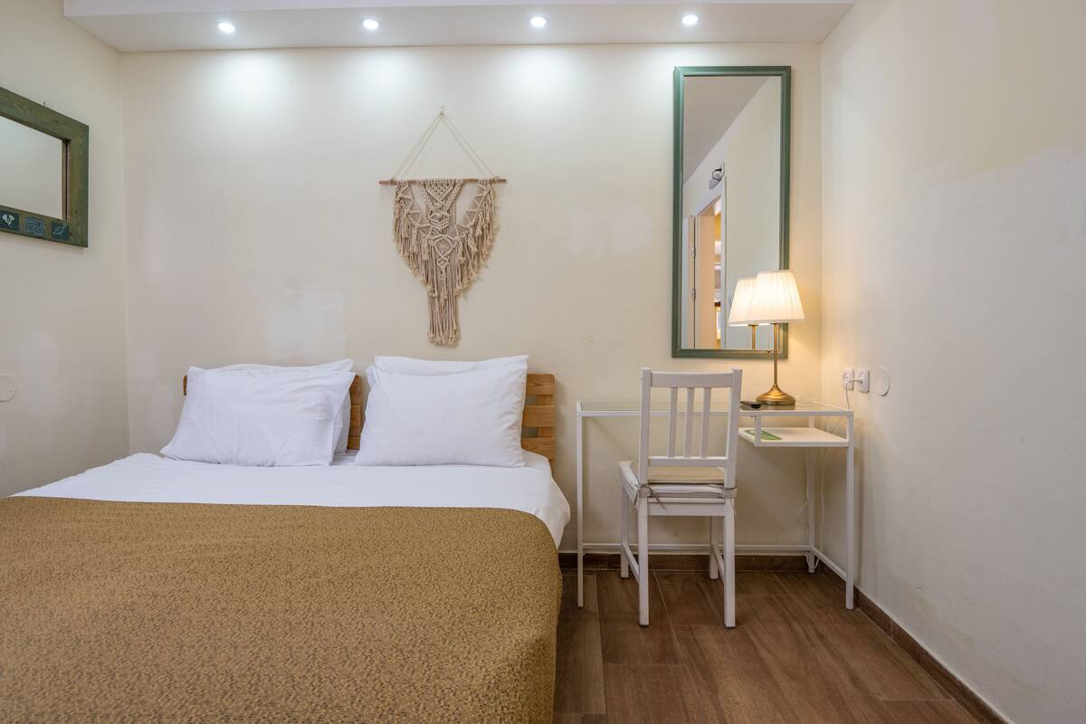 A hotel room with a bed, a desk, a chair, a lamp, and a macrame wall hanging.