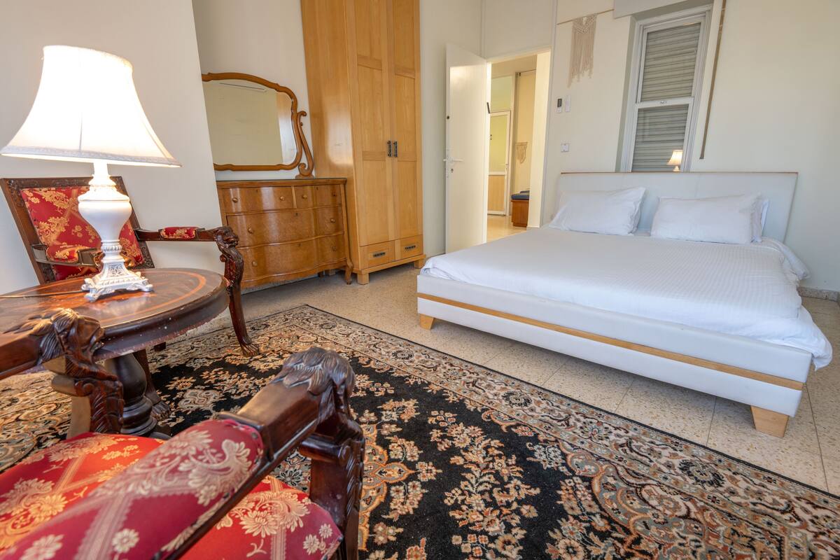 A bedroom with a bed, a wooden wardrobe, a mirror, a table with a lamp, and a red upholstered chair.
