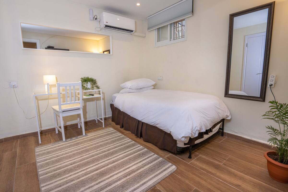 A bedroom with a bed, a desk, a chair, a mirror, and a potted plant.