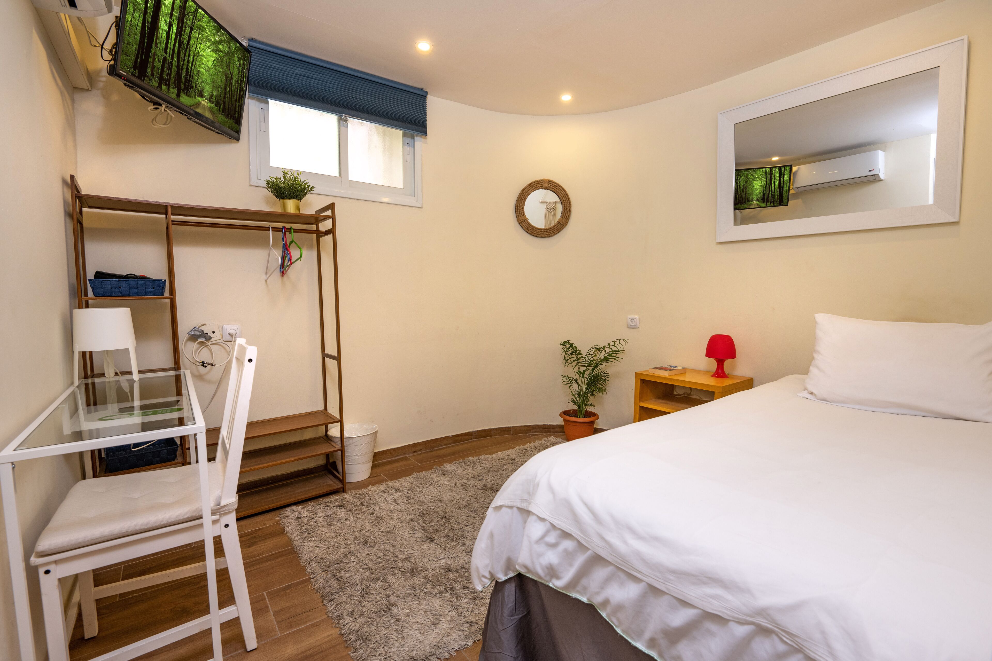 Un dormitorio con cama, escritorio, silla, mesita, planta en maceta, espejo y ventana con persianas.