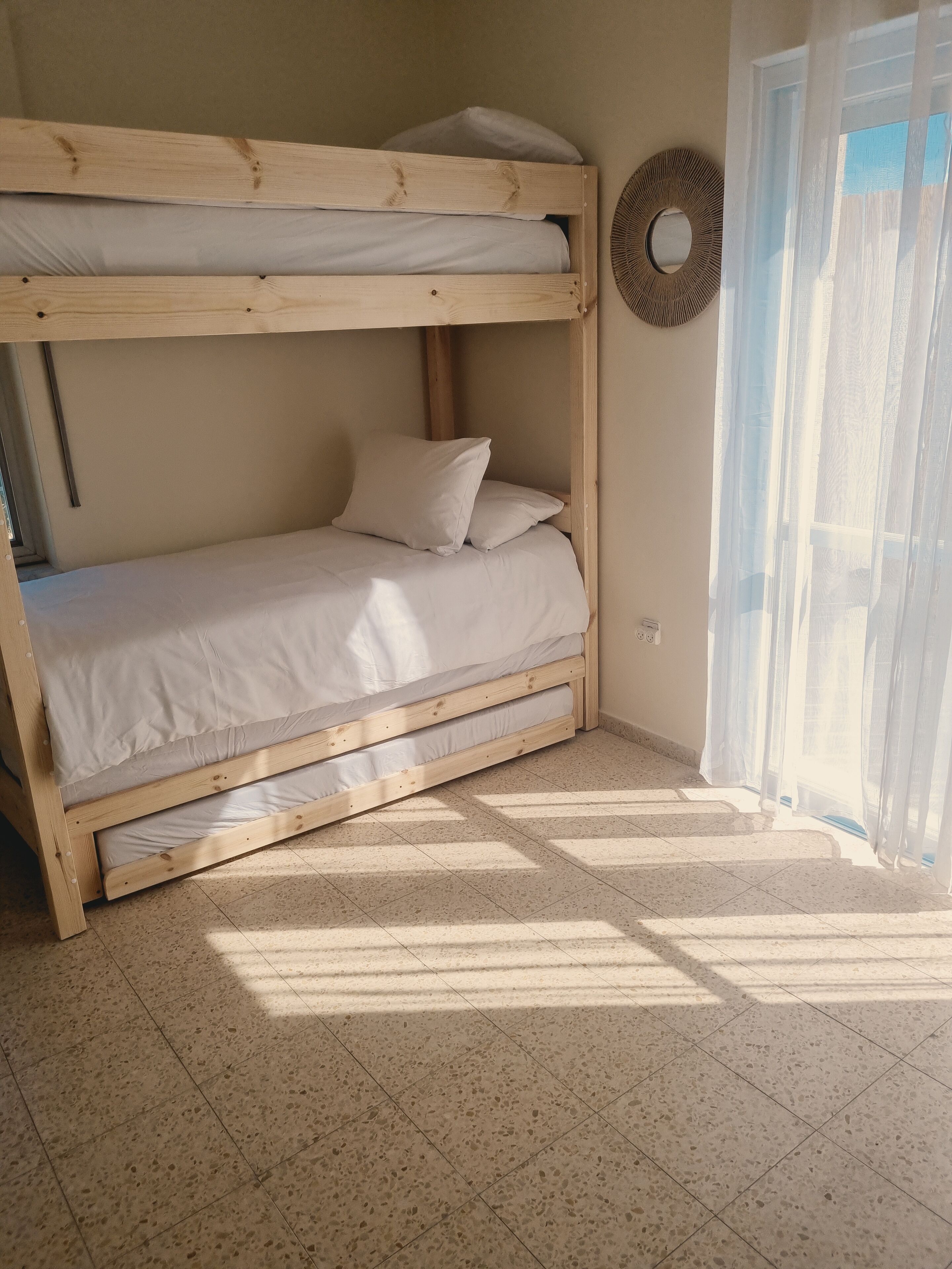 Una cama individual con ropa blanca, un espejo redondo en la pared y vistas al exterior a través de cortinas translúcidas.