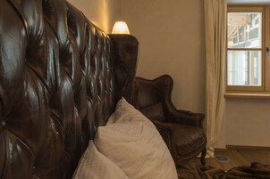 Un sofá de cuero marrón oscuro con respaldo capitonado, un sillón de madera y una ventana con cortinas color beige.