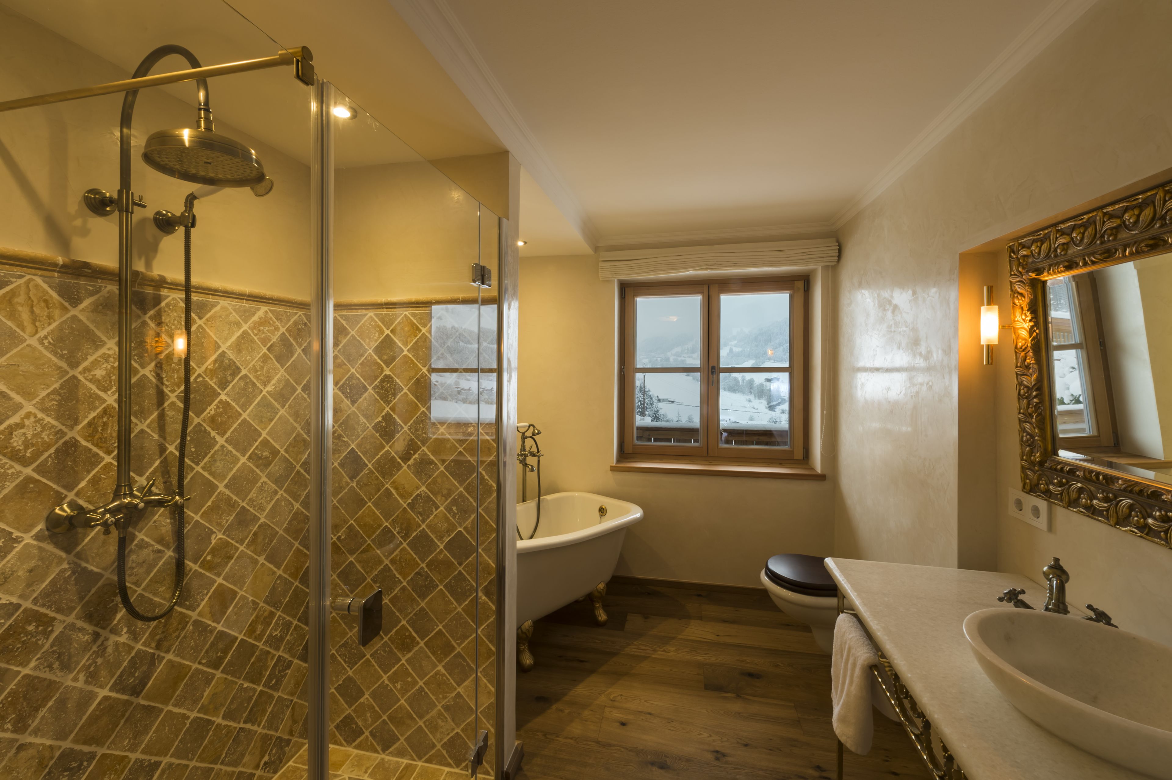 Un bagno con doccia vetro, vasca freestanding, lavabo e finestra vista montagne innevate.