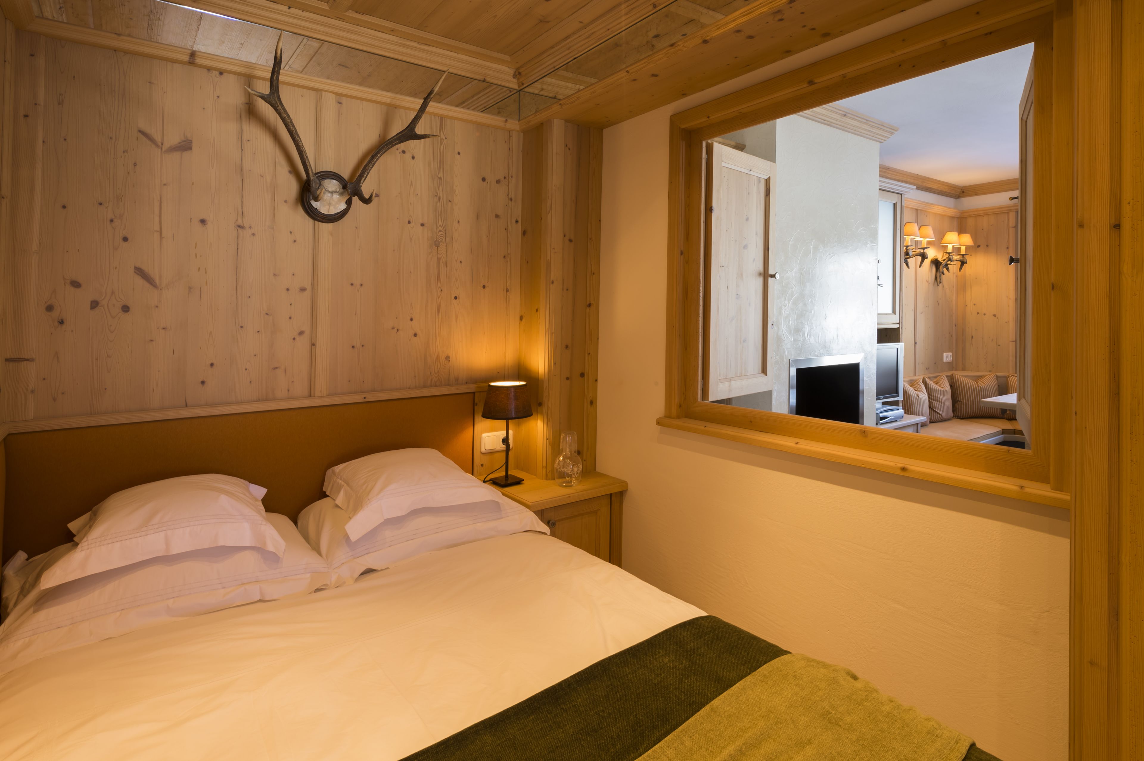 Ein Schlafzimmer mit einem Holzkopfende, einem Bett mit weißer Bettwäsche, einer Nachttischlampe und einem Fenster mit Blick auf einen anderen Raum.