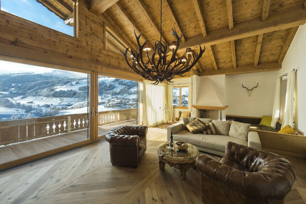 Ampla sala de estar com uma grande janela com vista para uma paisagem nevada, teto de madeira e um candelabro.