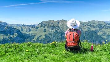 A person sitting on a grassy hill with a red backpack and a white hat, overlooking a mountainous landscape.