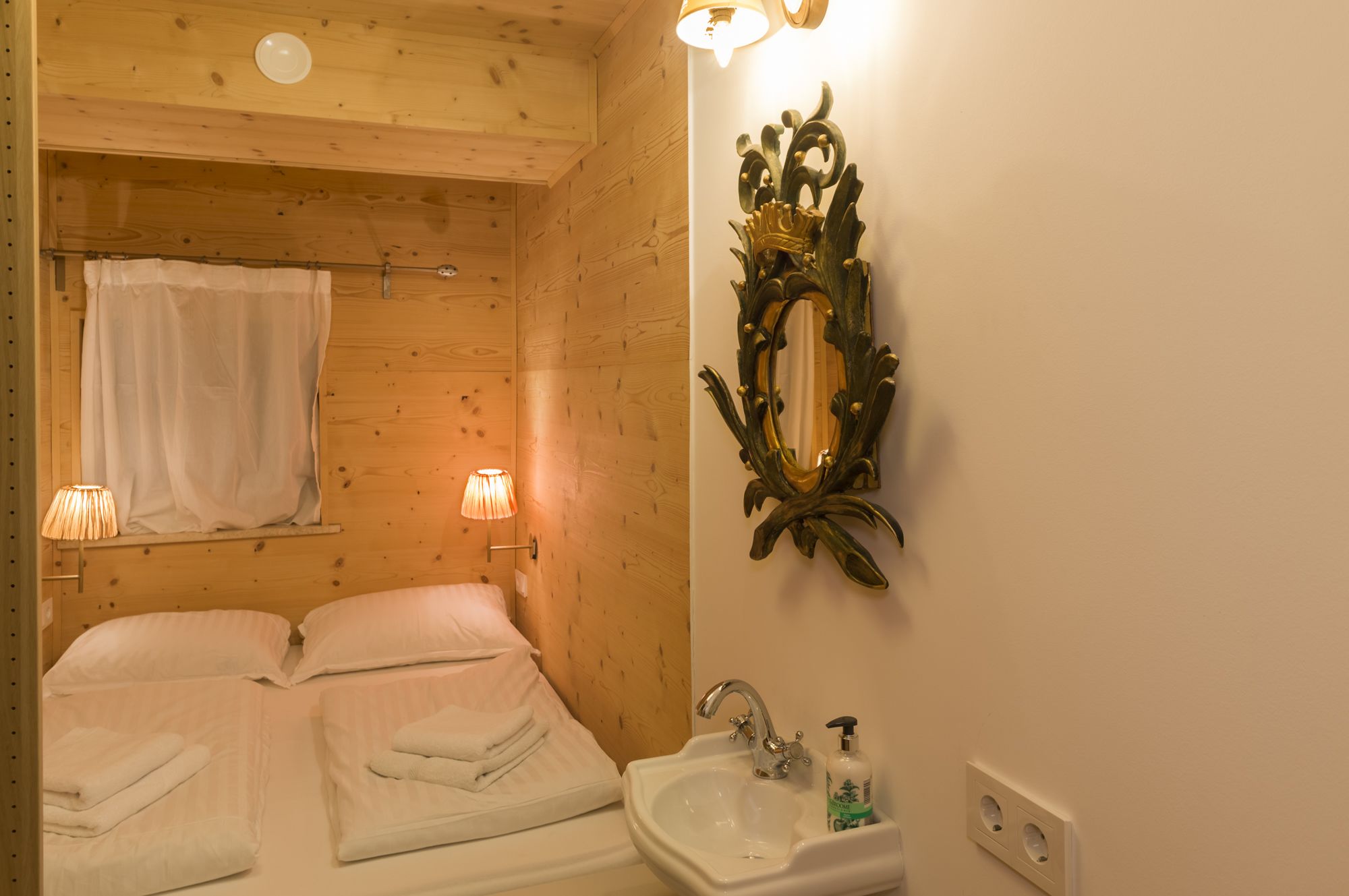 Ein holzgetäfeltes Zimmer im Chalet-Stil mit zwei Betten, einem Badezimmer mit Waschbecken und Spiegel sowie zwei Wandlampen.