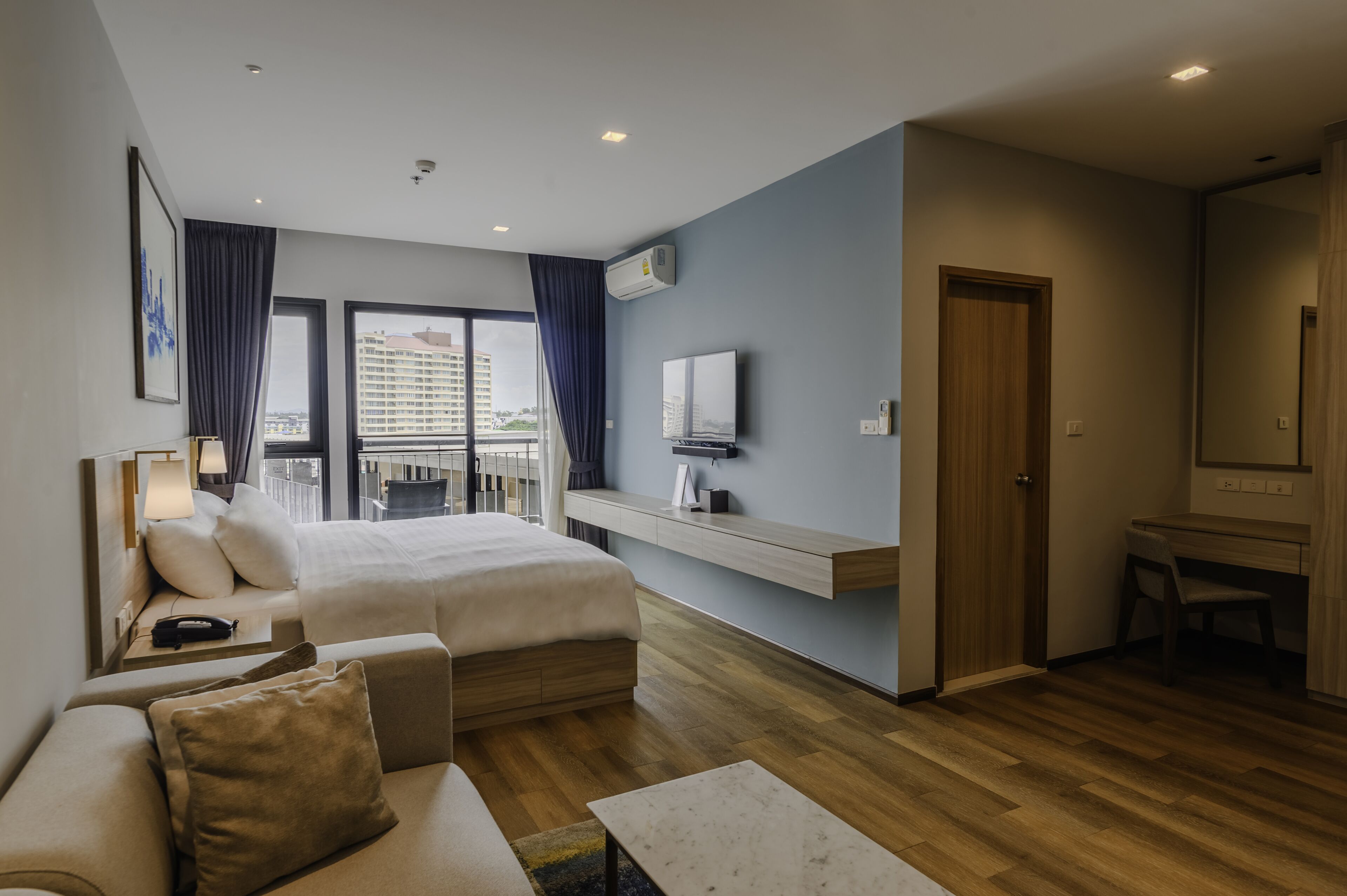 A modern hotel room with a bed, a sofa, a desk, and a balcony with a view of the city.