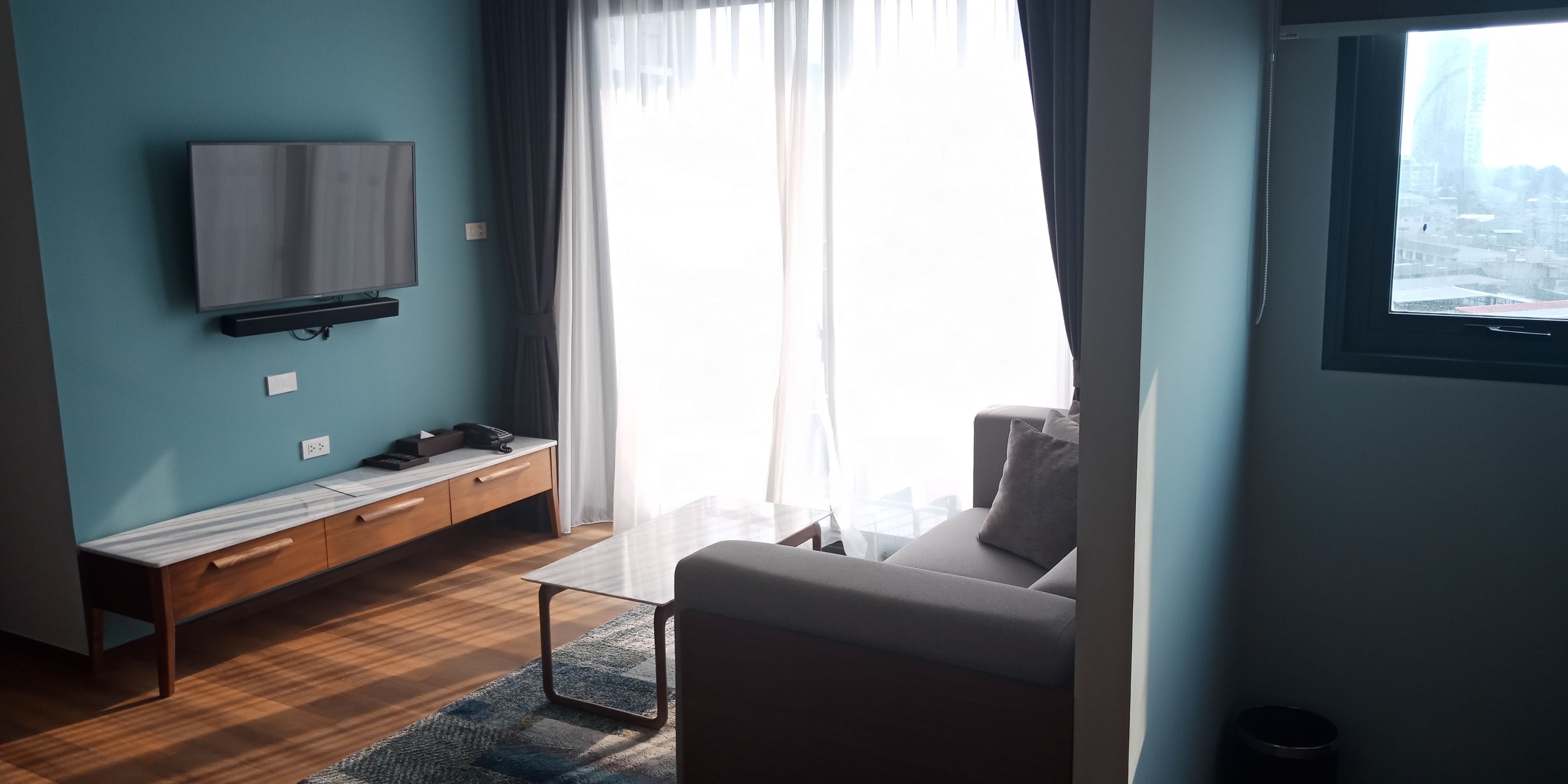 Een moderne woonkamer met een flatscreen televisie aan de muur, een houten bankje, een bank en een groot raam met dunne gordijnen.
