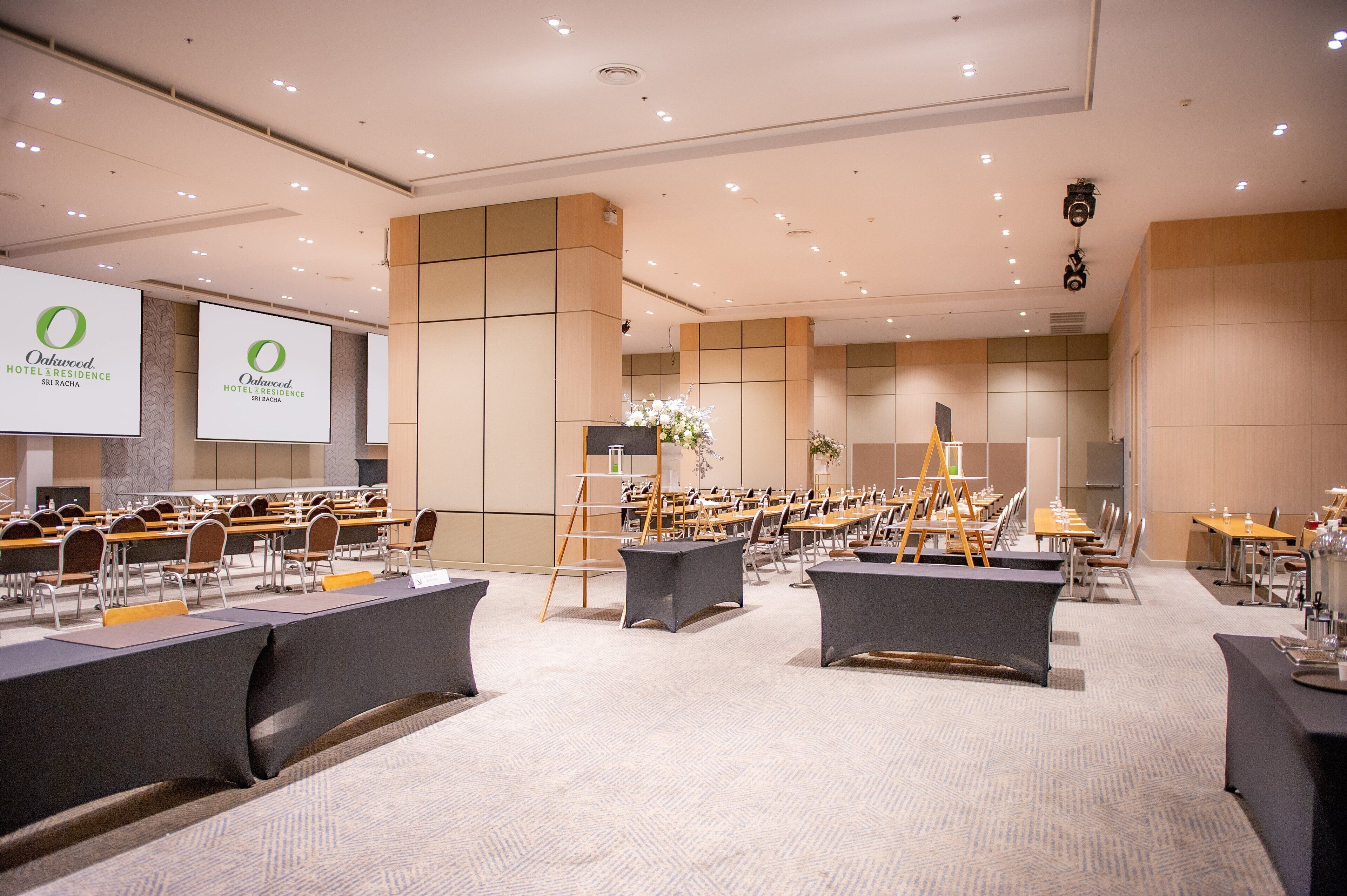 A conference room with multiple tables and chairs, a projector screen, and a large screen displaying the hotel name.