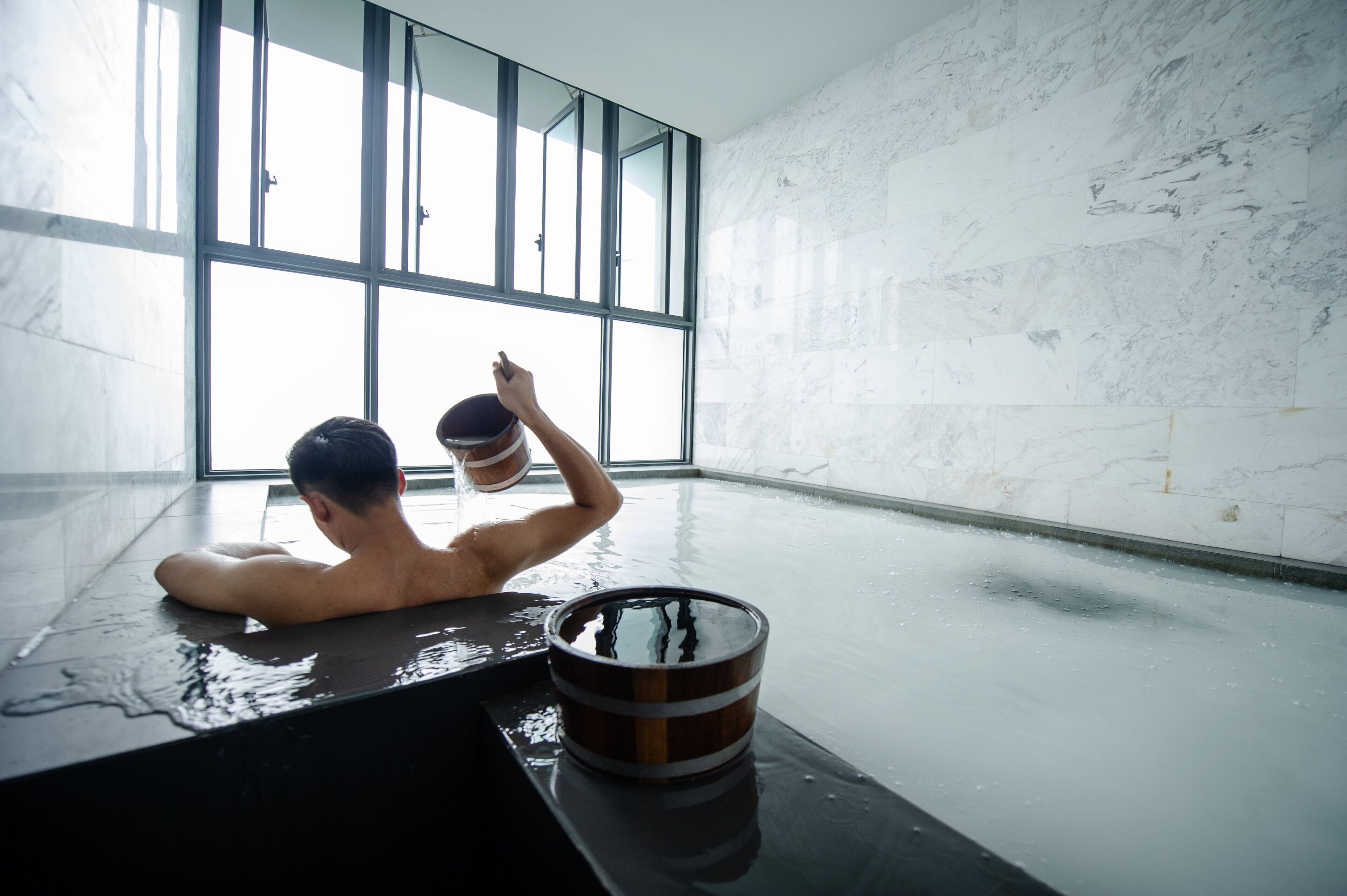 A person in a bathtub with a wooden bucket, large windows, and a marble wall.