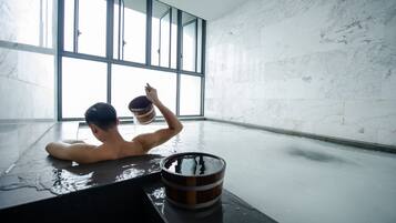 A person in a bathtub with a wooden bucket, large windows, and a marble wall.