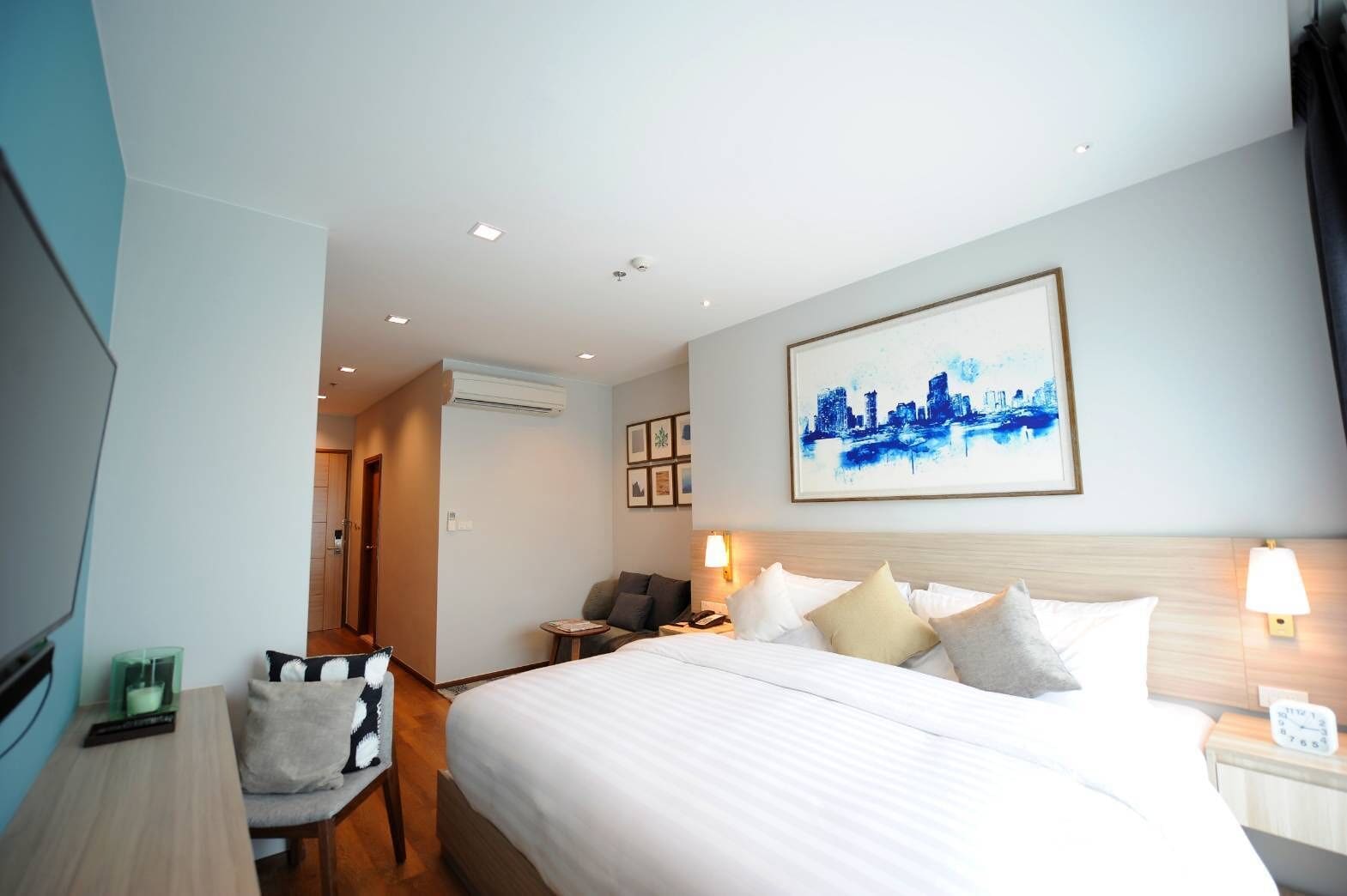 A hotel room with a large bed, a desk, a chair, and a cityscape painting on the wall.