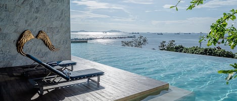 An infinity pool with a view of the sea and ships, lounge chairs, and a decorative wall.