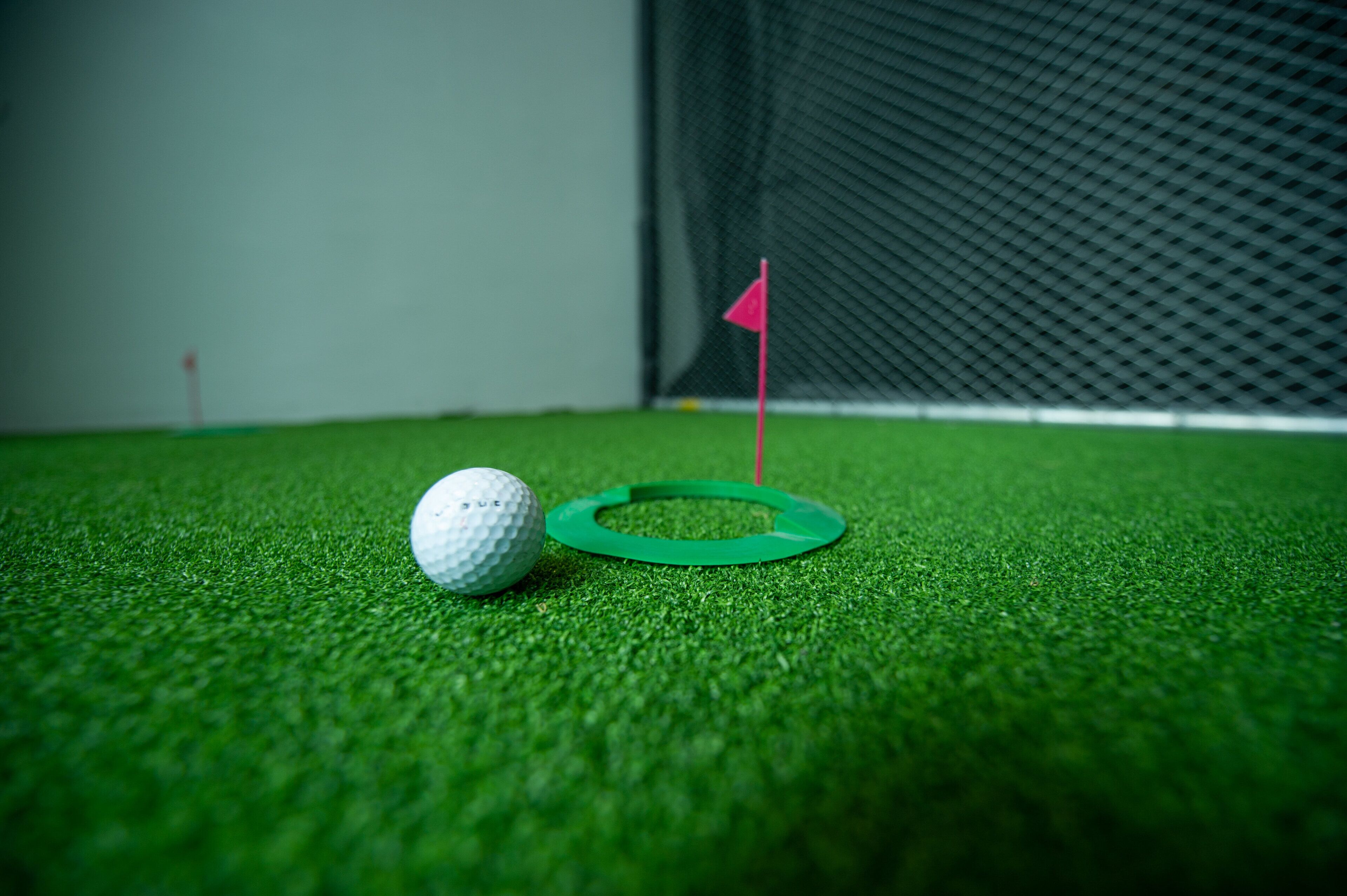 A golf ball on artificial grass near a green flag.