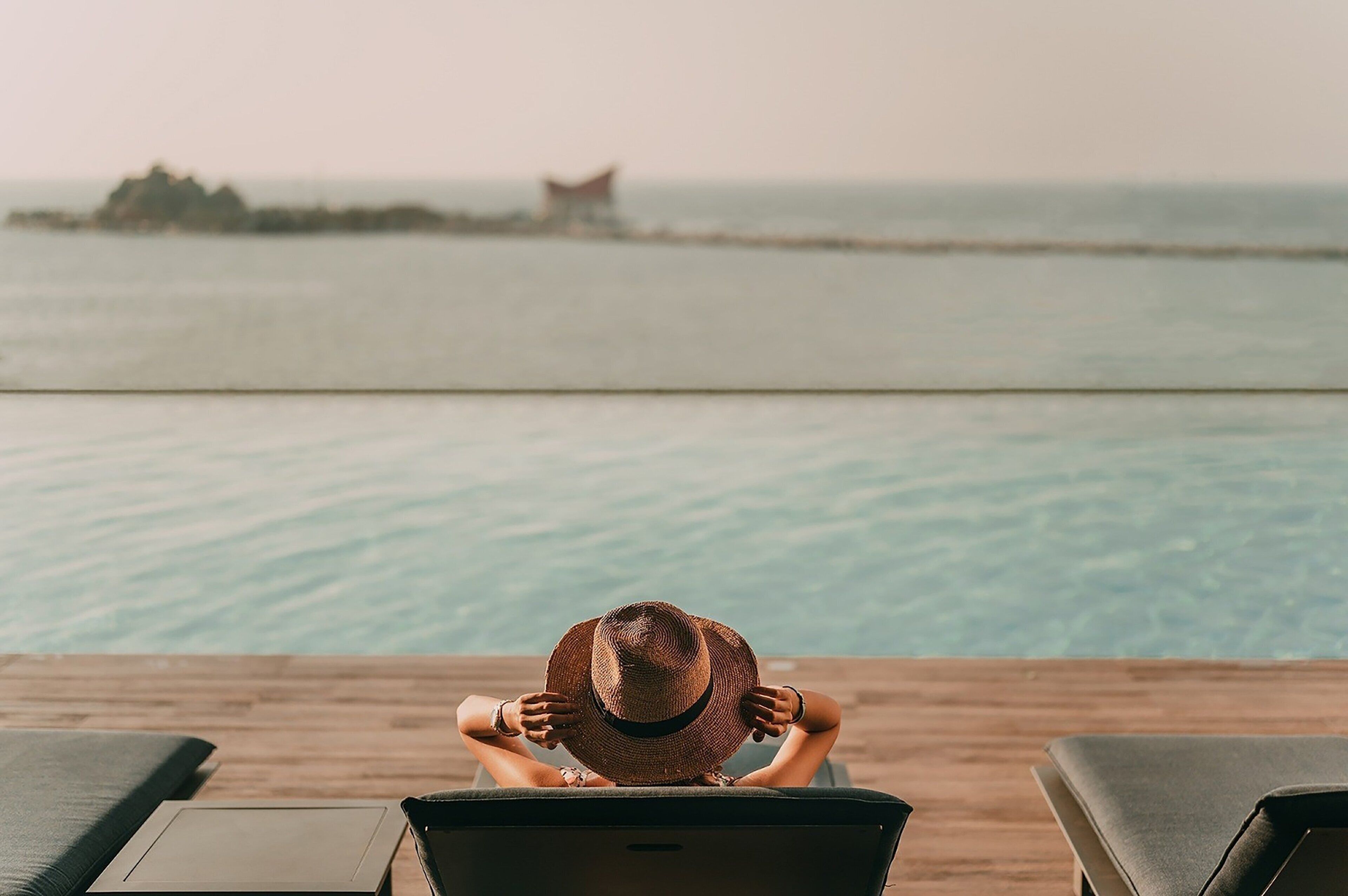 Eine Person sitzt am Pool auf einer Liege und hält einen Hut.