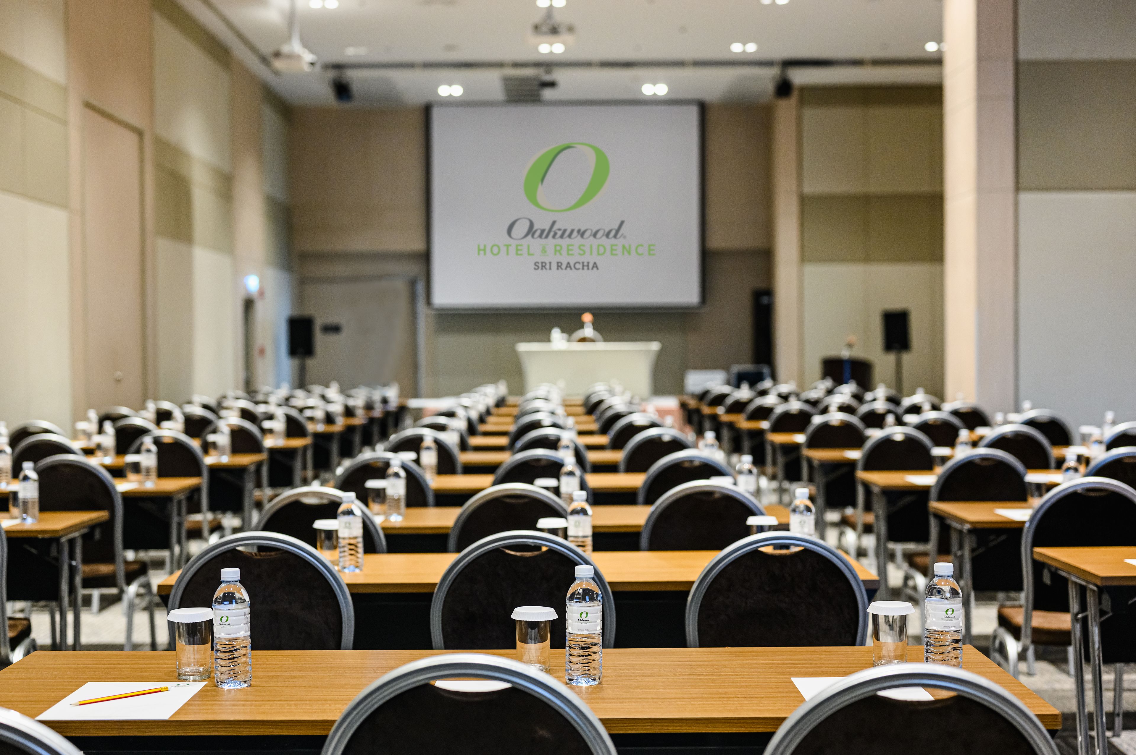 A conference room with rows of tables and chairs, a podium at the front, and a screen displaying the hotel name.