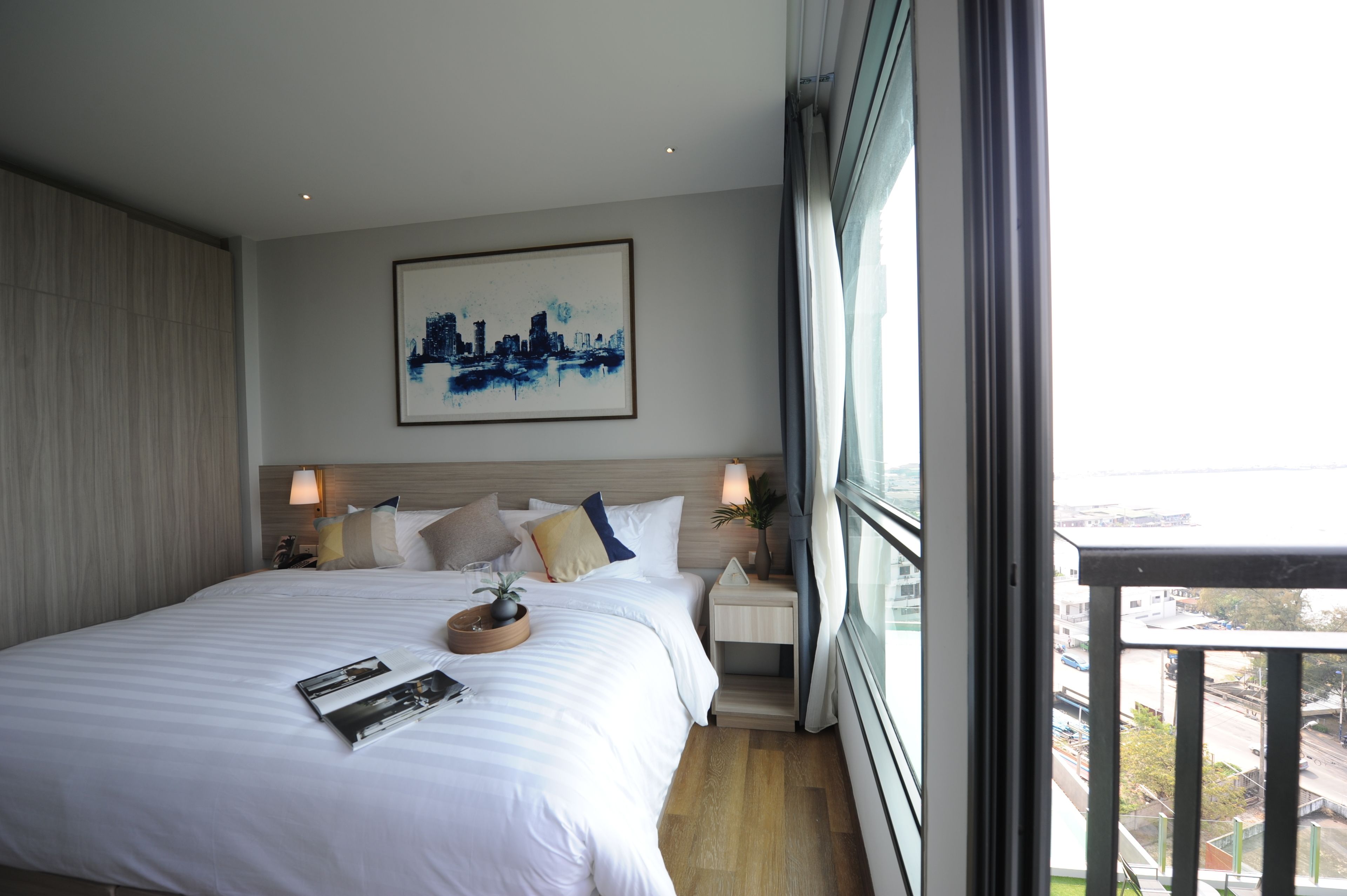 A modern hotel room with a large bed, two bedside tables, a cityscape painting, and a view of the city through the window.