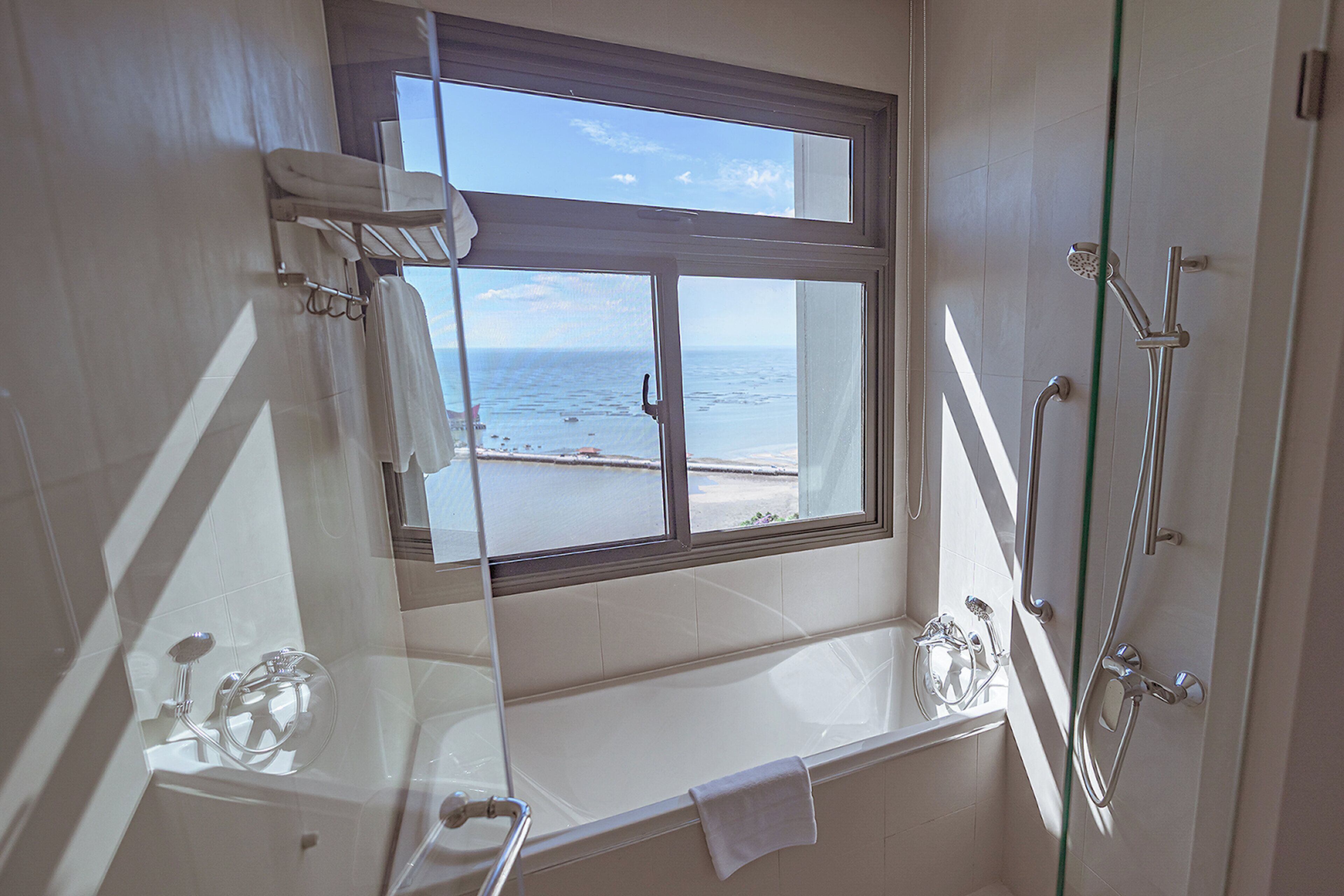 A bathroom with a bathtub, a shower, and a window overlooking the sea.