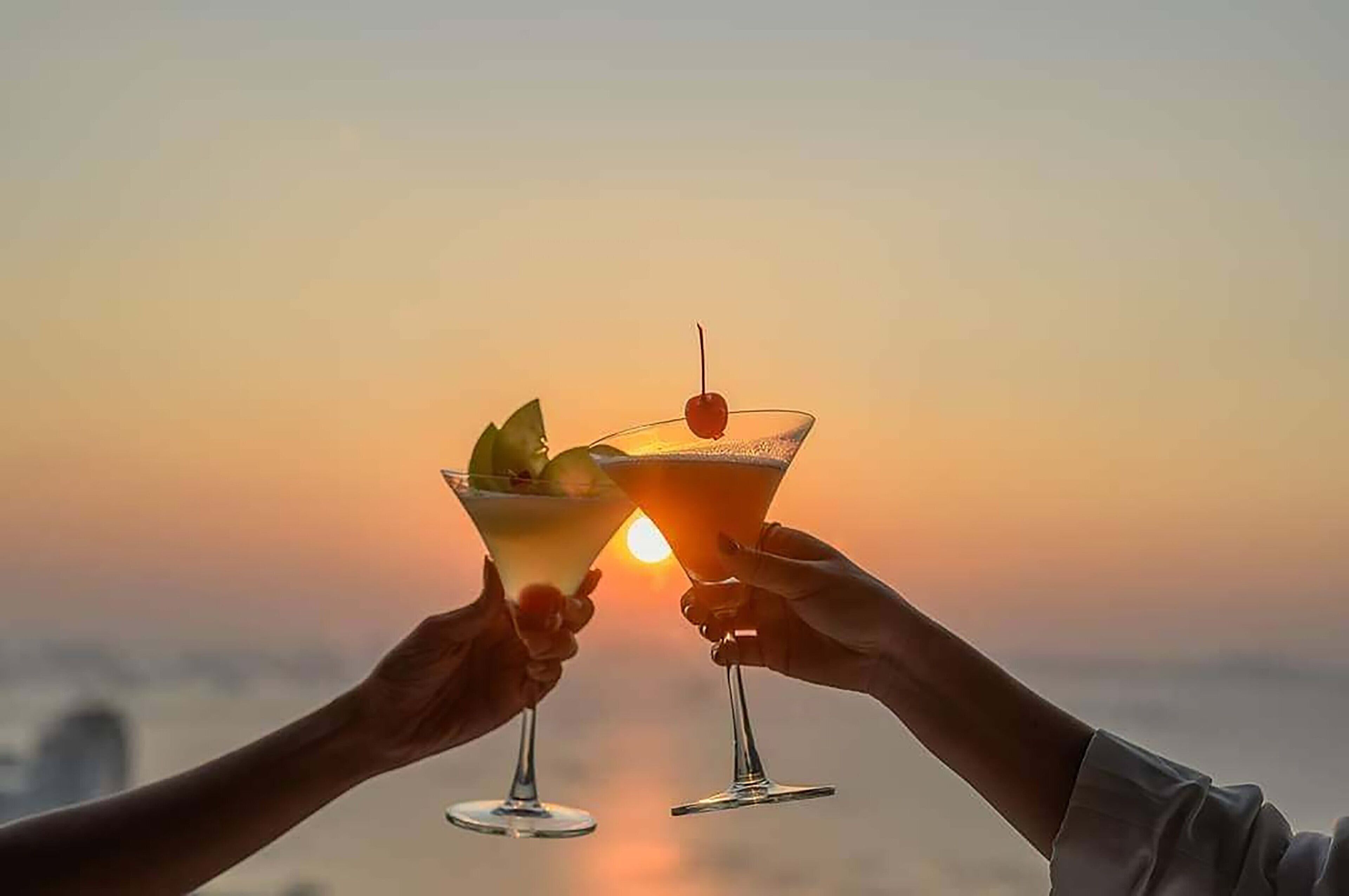 Two hands holding cocktail glasses with a sunset in the background.